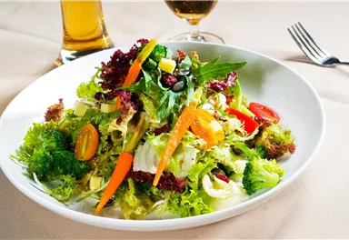A fresh salad with various types of leafy greens, tomatoes, carrots, and a colorful selection of other vegetables. In the background, there is a drink.