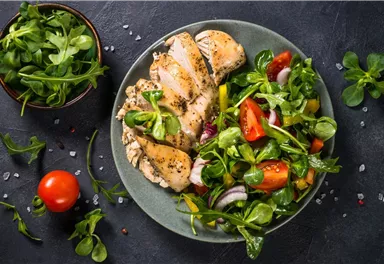 A plate with grilled chicken and fresh salad. Next to the plate is a bowl of arugula.
