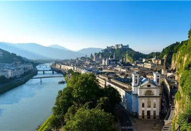 Eine malerische Aussicht auf die Stadt Salzburg mit dem Fluss Salzach im Vordergrund. Im Hintergrund erstrecken sich Berge und historische Gebäude.