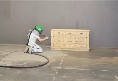 Eine Person in Schutzkleidung arbeitet an einem Holzschrank. Der Schrank steht auf einem unebenem Boden in einem offenen Raum.