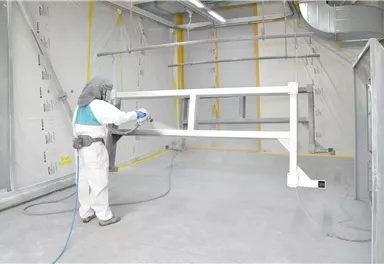 Ein Arbeiter in Schutzkleidung sprüht Farbe auf ein metallisches Gestell in einem sauberen, gut belüfteten Raum. Der Boden ist hell und die Wände sind grau mit gelben Markierungen.