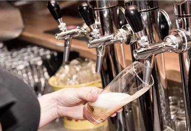 Eine Person zapft ein Bier aus einem Zapfhahn. Im Hintergrund sind Gläser und die Bar sichtbar.