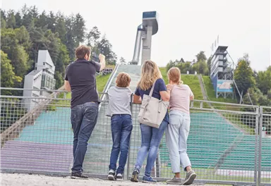 Eine Familie steht vor einer Sprungschanze im Freien. Im Hintergrund sind grüne Hügel und Sitzplätze zu sehen.