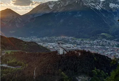 Eine malerische Aussicht auf eine Stadt umgeben von Bergen und Wald. Im Vordergrund ist eine Skisprungschanze zu sehen, die perfekt in die Landschaft integriert ist.