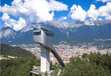 Ein modernes Aussichtsturm erhebt sich über der Stadt, umgeben von schönen Bergen und Bäumen. Ein strahlend blauer Himmel mit weißen Wolken vervollständigt die malerische Aussicht.