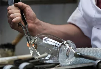 Ein Handwerker formt ein Glas mit einer Zange. Im Hintergrund sind Werkzeuge zur Glasbearbeitung sichtbar.