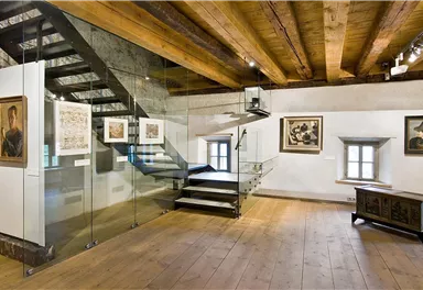 Ein moderner Ausstellungsraum mit einer gläsernen Treppe und Holzdecken. An den Wänden hängen mehrere gerahmte Bilder.