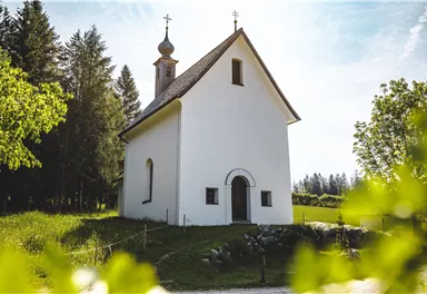 Eine kleine, weiße Kirche umgeben von Grün und Bäumen. Der Himmel ist blau mit wenigen Wolken.