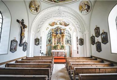 Eine helle Kirche mit Holzbankreihen und einem schönen Altar. An den Wänden hängen religiöse Bilder und ein Kreuz.