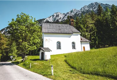 Eine kleine, weiße Kirche steht idyllisch in einer grünen Wiese. Im Hintergrund sind majestätische Berge zu sehen, und der Himmel ist klar.