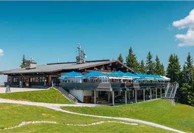 Ein modernes Restaurant in den Bergen mit einer großen Terrasse und blauen Sonnenschirmen. Umgeben von grünen Wiesen und Nadelbäumen unter einem klaren blauen Himmel.