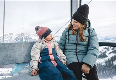 A mother and her child are sitting in a gondola overlooking snow-covered mountains. Both are wearing warm winter clothing and are smiling at each other.