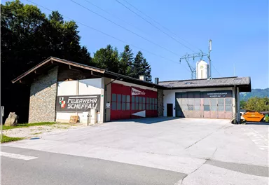 Ein Feuerwehrhaus in Scheffau mit einer roten Garagentür. Die Umgebung ist von Bäumen umgeben und der Himmel ist klar und blau.