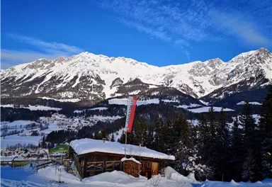 A picturesque mountain landscape with snow-covered peaks and a clear blue sky. In the foreground stands a small hut, surrounded by snow-covered trees.