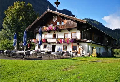 A traditional wooden house with colorful flowers in the windows. It is situated in a green setting with mountains in the background.