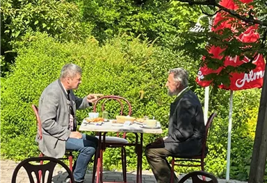 Two men are sitting at a table outdoors, enjoying a meal. Surrounded by lush greenery, they appear relaxed and engrossed in conversation.