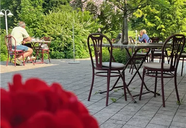 A cozy outdoor café with tables and chairs. In the foreground, red and pink flowers are blooming, while in the background, guests are relaxing.