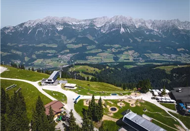 A picturesque mountain landscape with green meadows and snow-capped peaks. In the foreground, recreational areas and buildings can be seen.