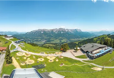 A picturesque mountain landscape with green meadows and snow-covered peaks in the background. In the foreground, playgrounds and buildings can be seen that offer a beautiful view.