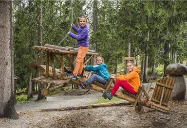 Drei Kinder spielen auf einer begehbaren Holzspielzeugmaschine im Wald. Sie haben bunte Jacken an und lachen fröhlich.
