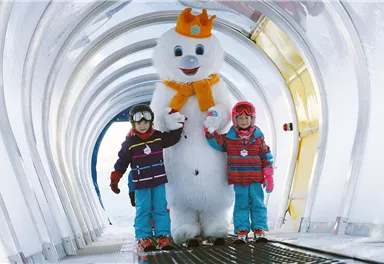Two children are standing next to a large snowman in a winter alley. They are wearing colorful winter clothes and smiling happily.