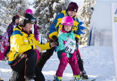 A happy child is learning to ski with the help of a trainer. They are standing at the starting line in a snowy landscape.