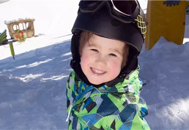 A smiling child in the snow is wearing a helmet and a colorful jacket. In the background, playground equipment and the snow-covered landscape can be seen.