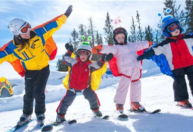 A ski instructor and three children are standing on skis in the snow. They are all wearing colorful ski clothes and helmets while posing happily.