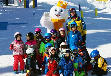 A group of children in colorful ski suits is standing in the snow and smiling. In the background, there is a large, friendly snowman wearing a crown.