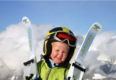 A smiling child is wearing a helmet and holding skis in their hands. In the background are snow-covered mountains and a blue sky.