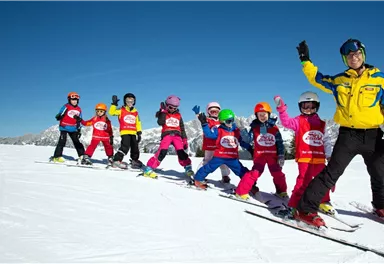 A group of children is standing on skis in the snow and smiling. A ski instructor is waving to them next to the group.
