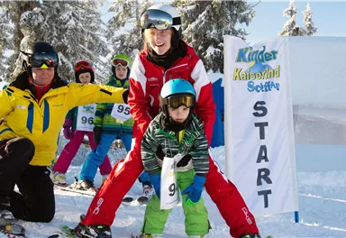A cheerful ski race for children with two teachers and several small skiers. The start banner is clearly visible in the background.
