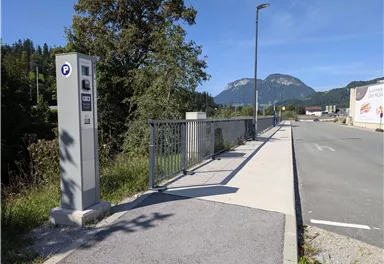 A parking lot with a parking meter is located on a quiet street. In the background, green mountains and a clear sky can be seen.
