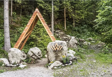 A wooden triangular sign stands between trees and large stones. The path leads through a green, wooded landscape.