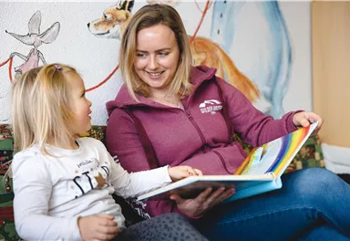 A woman is reading a book to a little girl. Colorful murals can be seen in the background.