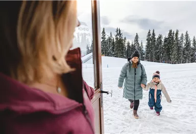A mother and her child happily come out of the house into the snowy landscape. In the background, there are tall trees and a gray sky.