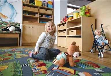A cheerful little girl is sitting on a colorful carpet in a children's room. Around her are toys and furniture for children.