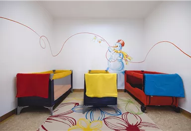 A friendly room with three baby cribs in bright colors. On the wall, there is a cheerful snowman depicted with a colorful string.