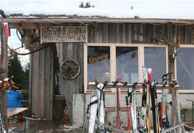 A rustic ski hut with the sign "STALL-BAR". In the foreground, skis are standing on a wooden rack.