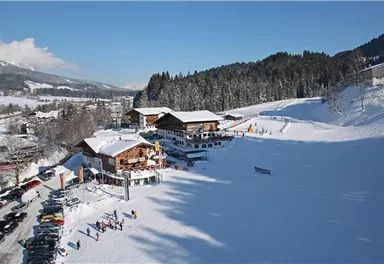 A picturesque mountain landscape with snow-covered hills and traditional huts. In the foreground, skiers and cars are visible in a parking lot.