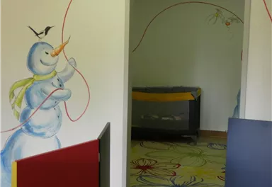 A friendly snowman with a red scarf stands against a wall while a fox sits above it. The image depicts an inviting room with colorful furniture and a cheerful floor pattern.