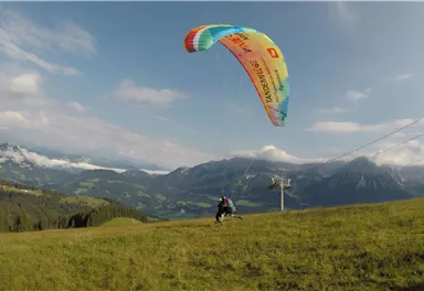 Eine Person fliegt mit einem Gleitschirm über eine grüne Wiese und beeindruckende Berglandschaft. Der Himmel ist blau mit einigen Wolken.