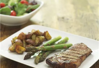 A plate with grilled steak, green beans, and fried potatoes. In the background, there is a bowl of fresh salad.