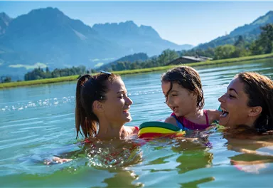 Ein sonniger Tag am Wasser mit zwei Frauen und einem Kind. Sie haben Spaß beim Spielen im Wasser mit einem bunten Ball und einer schönen Berglandschaft im Hintergrund.