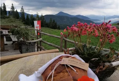Ein frisch gebackenes Brot auf einem Tisch, umgeben von bunten Blumen. Im Hintergrund sind sanfte Hügel und ein bewölkter Himmel zu sehen.