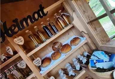 Ein Holzregal voller hausgemachter Produkte, darunter Flaschen mit verschiedenen Flüssigkeiten und frisch gebackene Brötchen. Der Raum strahlt eine einladende, rustikale Atmosphäre aus.