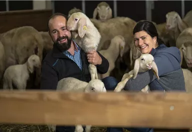 Ein Mann und eine Frau halten jeweils ein Lamm in der Hand, umgeben von weiteren Schafen in einem Stall. Beide lächeln freundlich in die Kamera.