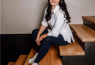 A smiling woman is sitting on a staircase made of wood and metal. She is wearing a white shirt and looking at the camera.