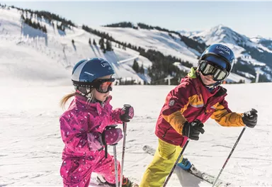Two children are skiing on a snow-covered slope. In the background, there are snow-covered mountains and a clear blue sky.