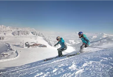 Two skiers on a snow-covered slope with mountains in the background. The sky is clear and the landscape is winterly beautiful.